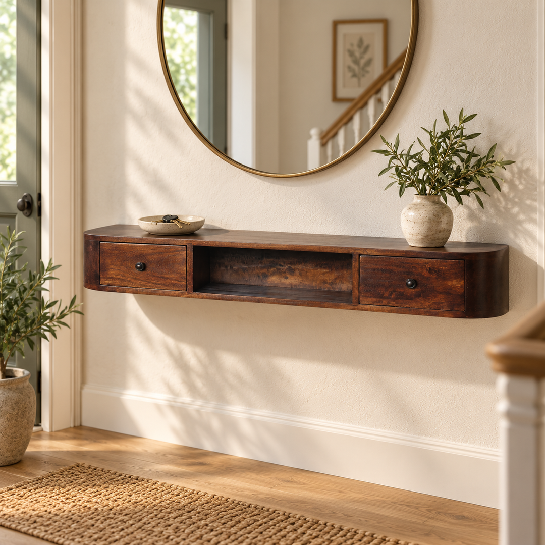 A floating dark wood console shelf mounted on a cream wall in a bright entryway, styled with a round gold-framed mirror, a small bowl, and potted green plants.